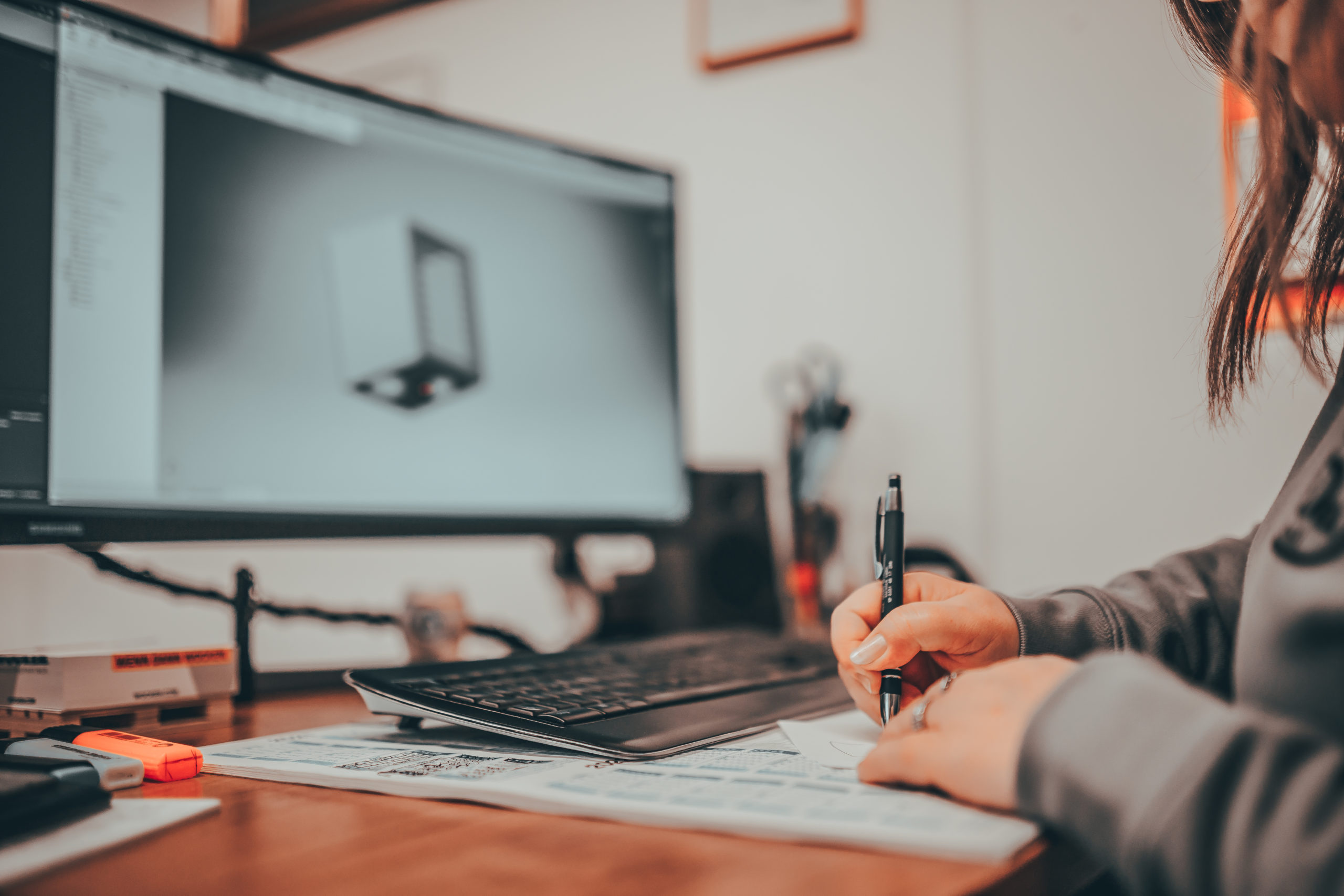 Frau schreibt am Schreibtisch etwas auf Papier. Im Hintergund ist ein Büro mit Monitor auf der eine CAD-Software geöffnet ist.